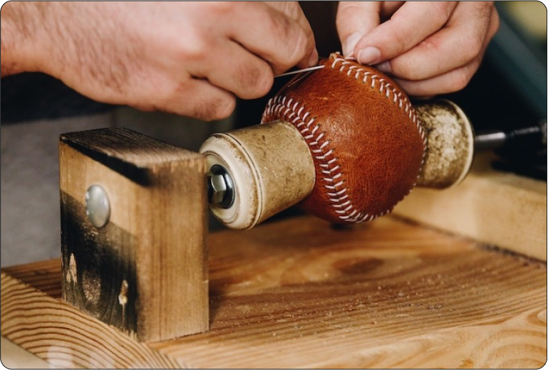 Hands carefully hand-stitching a Glove Tan Yardball leather ball on a wooden stitching machine, showcasing handcrafted artisan production in Kansas City